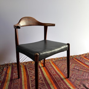 May include: A wooden chair with a dark brown finish and a curved backrest that extends into armrests. The seat is upholstered in black material. The chair features four tapered legs and a smooth, polished wood grain texture throughout the frame.