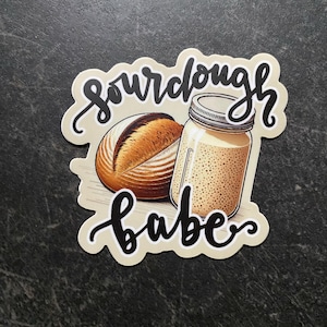 May include: A sticker with a sourdough bread loaf and a jar of sourdough starter. The sticker has the text "sourdough babe" in black cursive.