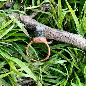 Puede incluir: Un anillo de cobre con una montura de piedras preciosas verdes y azules apiladas. El anillo está sobre un lecho de hierba verde.