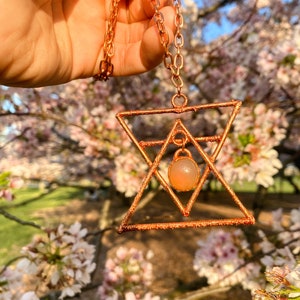 May include: A copper wire necklace with a geometric pendant featuring a peach-coloured gemstone. The pendant is in the shape of a star with a triangle inside.