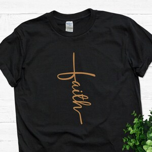 Puede incluir: Camiseta negra con una cruz marr&oacute;n y la palabra "faith" escrita verticalmente en la cruz.