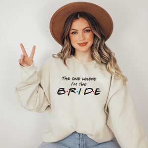 May include: A beige sweatshirt with the text "The One Where I'm The B.R.I.D.E." in black with colourful dots. The sweatshirt is worn by a person with long brown hair and a brown hat.
