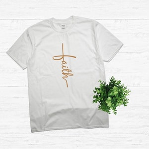 Puede incluir: Camiseta blanca con una cruz marr&oacute;n y la palabra "faith" escrita verticalmente.