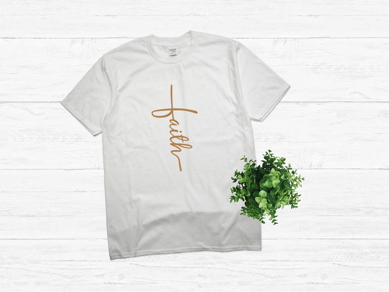 Puede incluir: Camiseta blanca con una cruz marr&oacute;n y la palabra "faith" escrita verticalmente.