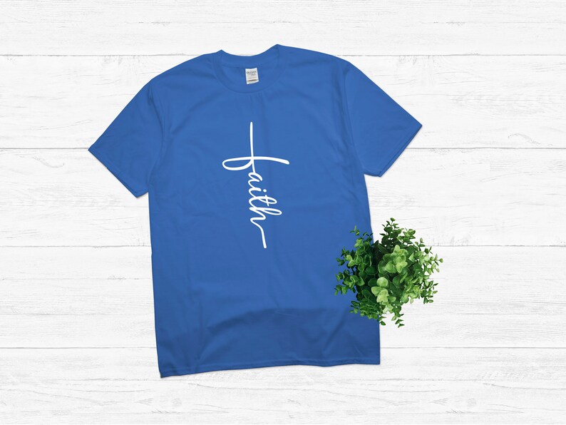 Puede incluir: Una camiseta azul real con una cruz blanca y la palabra "faith" escrita verticalmente en ella.