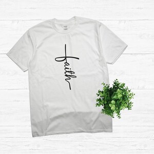 Puede incluir: Camiseta blanca con una cruz negra y la palabra "faith" escrita verticalmente en la cruz.