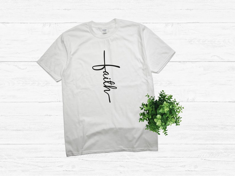 Puede incluir: Camiseta blanca con una cruz negra y la palabra "faith" escrita verticalmente en la cruz.