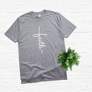 Puede incluir: Una camiseta gris jaspeada con una cruz blanca y la palabra "faith" escrita verticalmente en ella.