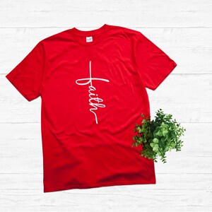 Puede incluir: Camiseta roja con una cruz blanca y la palabra "faith" escrita verticalmente en ella.
