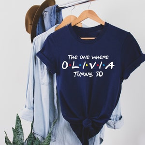 May include: A navy blue t-shirt with the text "The One Where Olivia Turns 30" in white with colorful dots. The text is styled like the Friends logo.
