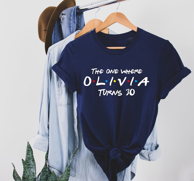 May include: A navy blue t-shirt with the text "The One Where Olivia Turns 30" in white with colorful dots. The text is styled like the Friends logo.