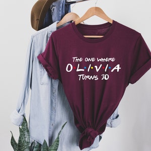 May include: Burgundy t-shirt with white text that reads "The One Where Olivia Turns 30". The text is styled like the Friends logo with colorful dots.