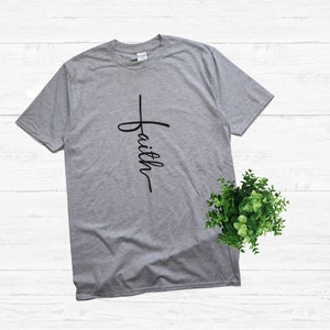 Puede incluir: Una camiseta gris con una cruz negra y la palabra "faith" escrita verticalmente en ella.
