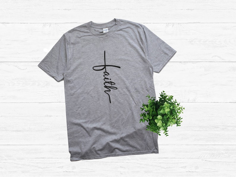 Puede incluir: Una camiseta gris con una cruz negra y la palabra "faith" escrita verticalmente en ella.