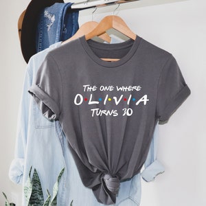 May include: Gray t-shirt with the text "The One Where Olivia Turns 30" in a white font. The text is styled to look like the title card from the TV show Friends.