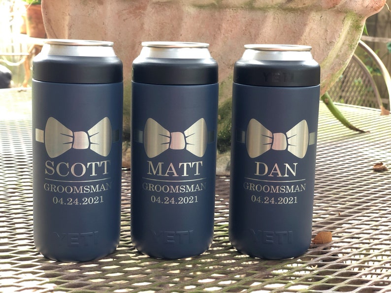 May include: Three blue Yeti Rambler 12 oz.  cans with a silver bow tie design and personalized text. The text reads "Scott Groomsman 04.24.2021", "Matt Groomsman 04.24.2021", and "Dan Groomsman 04.24.2021".