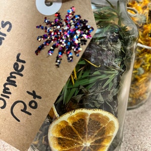 May include: A glass jar containing dried herbs, a dried orange slice, and other botanicals. A brown tag with the words "Simmer Pot" is attached with twine. The tag is decorated with a glittery starburst in red, white, and blue.