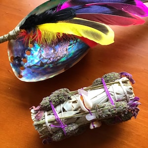 May include: A white sage smudge stick wrapped with purple string and lavender buds. The smudge stick is tied with white string and includes small white seashells. The smudge stick is sitting on a wooden surface.