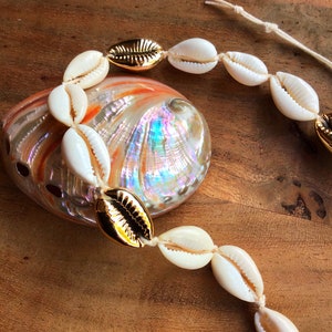 May include: A white shell necklace with gold accents. The necklace is made of cowrie shells and is strung on a white cord. The necklace is lying on a wooden surface.