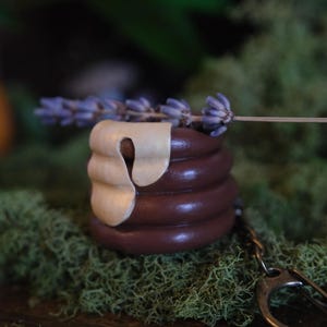 May include: A brown, beehive-shaped keychain with a cream-colored accent and a sprig of lavender. The keychain is made of a dark brown material and sits on a bed of green moss. A silver-colored keyring is attached.
