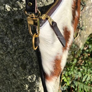 May include: A close-up of a handbag with a cowhide pattern in brown and white. The bag features a gold zipper and hardware, with a black leather strap and trim. The bag is set against a natural, textured background.