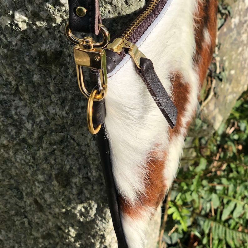 May include: A close-up of a handbag with a cowhide pattern in brown and white. The bag features a gold zipper and hardware, with a black leather strap and trim. The bag is set against a natural, textured background.