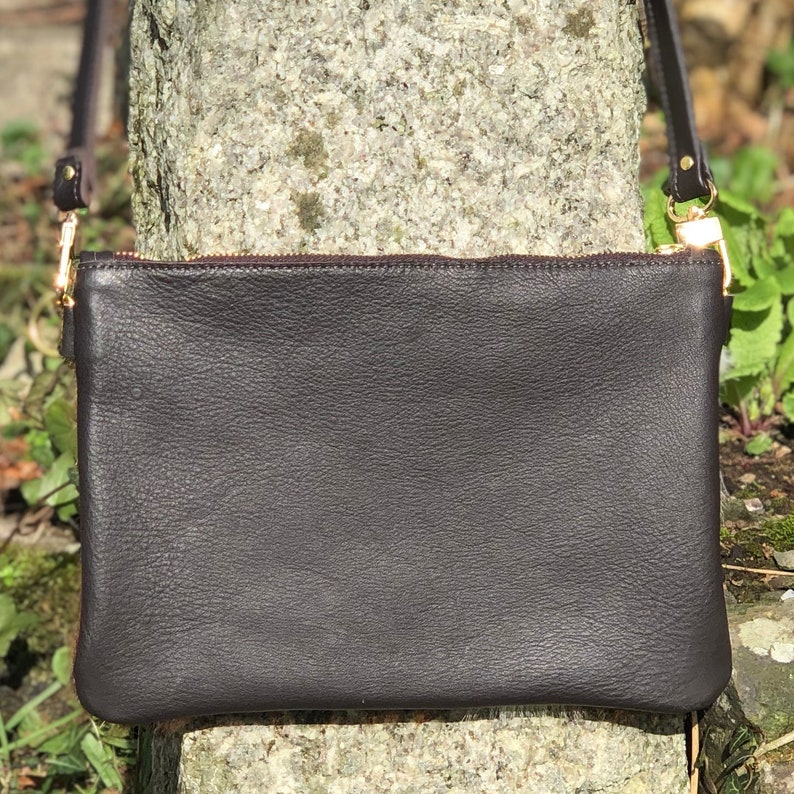 May include: A dark brown leather crossbody bag with a gold zipper and strap attachments. The bag is rectangular and has a textured surface. The strap is thin and dark brown.