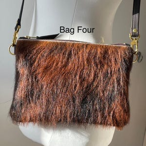 May include: A rectangular crossbody bag with a brown and black fur-like texture. The bag has a zipper closure and gold-colored hardware. The text "Bag Four" is printed on the top of the bag. The bag has a black strap.