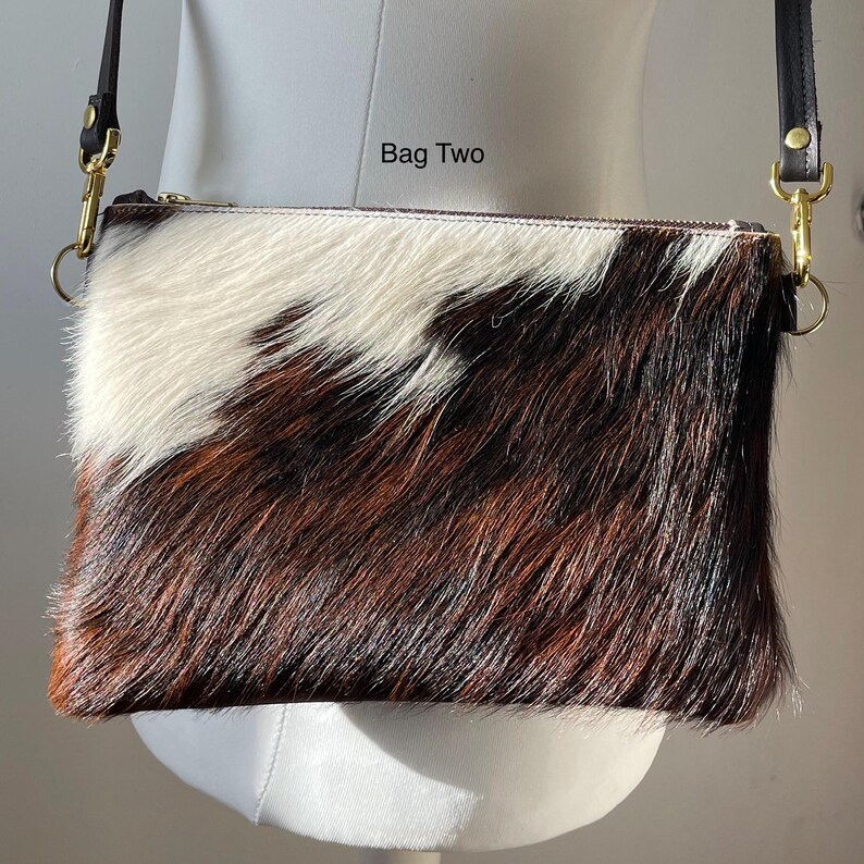 May include: A rectangular crossbody bag with a cowhide pattern. The bag features a mix of white, brown, and black fur. It has a gold zipper and black leather straps with gold hardware. The text "Bag Two" is visible on the bag.