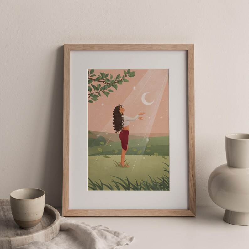 Puede incluir: Una impresi&oacute;n art&iacute;stica enmarcada representa a una persona de pie en un campo, con los brazos extendidos hacia una luna creciente y rayos de luz. La persona lleva una parte superior blanca y pantalones rojos. El fondo presenta un cielo rosa, colinas verdes y una rama de &aacute;rbol.