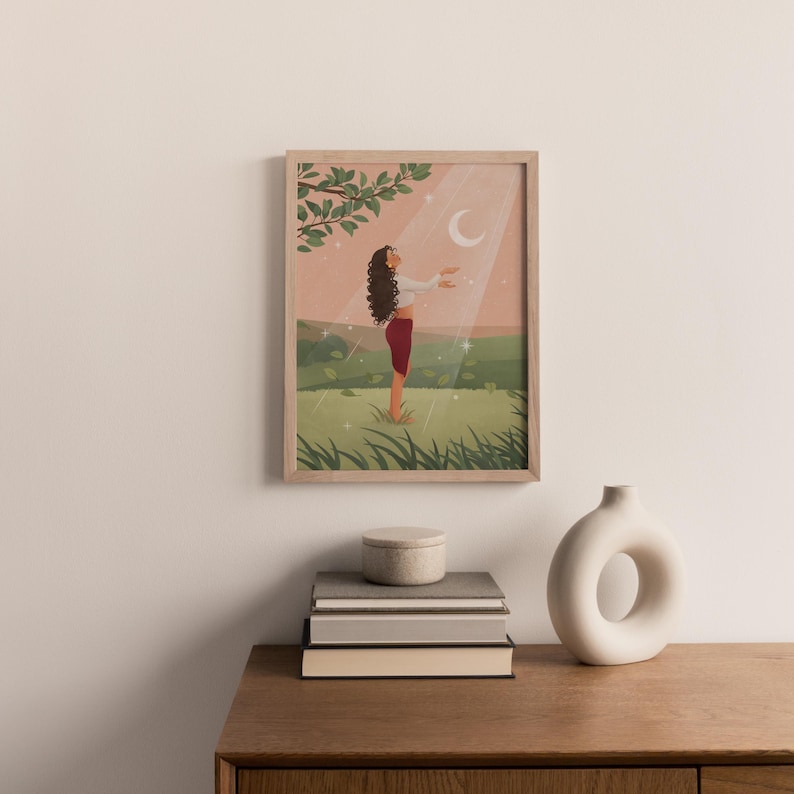 Puede incluir: Una impresi&oacute;n art&iacute;stica enmarcada representa a una persona en un campo, extendiendo la mano hacia una luna creciente. La obra de arte presenta una paleta de colores rosa, verde y blanco, con un rayo de luz. La impresi&oacute;n se exhibe sobre una mesa de madera con libros y un jarr&oacute;n.