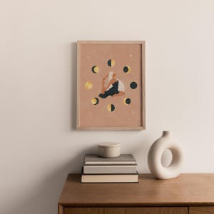 May include: A framed art print featuring a woman in a yoga pose surrounded by moon phases. The artwork has a warm, neutral background with gold, black, and white celestial elements. The frame is a light wood color, and the print is displayed on a wooden surface.