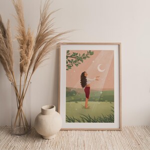 Puede incluir: Una impresi&oacute;n art&iacute;stica enmarcada representa a una persona en un campo, con los brazos extendidos hacia una luna creciente. La persona lleva una blusa blanca y una falda roja. La obra de arte tiene una paleta de colores suaves con un marco de madera marr&oacute;n claro.