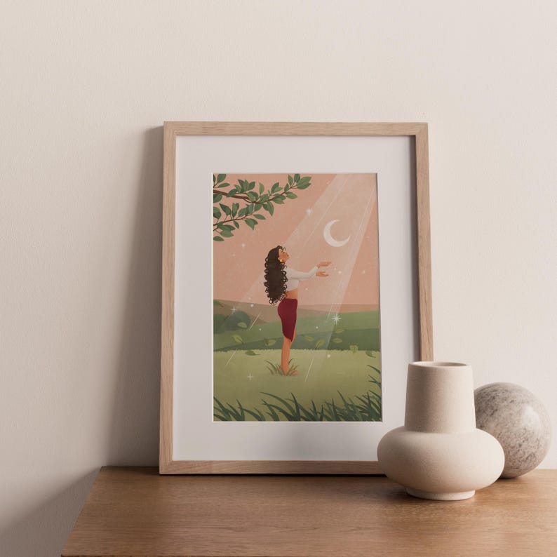 Puede incluir: Impresi&oacute;n art&iacute;stica enmarcada que representa a una mujer de pie en un campo, extendiendo la mano hacia una luna creciente en un rayo de luz. La obra de arte tiene una paleta de colores suaves con tonos verdes, rosas y blancos. La impresi&oacute;n est&aacute; en un marco de madera clara.