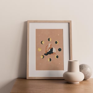 May include: A framed art print featuring a woman in a yoga pose surrounded by moon phases. The artwork has a warm, earthy color palette with gold and black accents. The print is displayed on a wooden surface, with a vase and decorative sphere in the foreground.