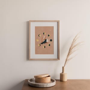 May include: Framed art print featuring a person in a yoga pose surrounded by moon phases. The artwork has a warm color palette with gold and black accents. The print is displayed in a light wood frame, adding a touch of natural elegance to the decor.