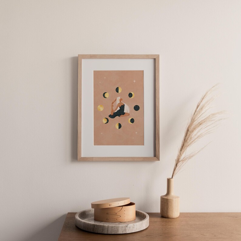 May include: Framed art print featuring a person in a yoga pose surrounded by moon phases. The artwork has a warm color palette with gold and black accents. The print is displayed in a light wood frame, adding a touch of natural elegance to the decor.