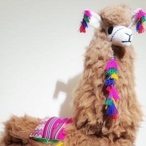 May include: A brown plush llama toy with a colorful saddle and tassels. The llama has a white face with black eyes and a pink nose.