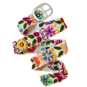 May include: A white embroidered belt with a silver buckle. The belt is adorned with colorful floral patterns in shades of pink, yellow, blue, and orange, with green leaves. The belt is approximately 1.5 inches wide.