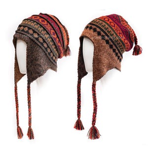 Könnte beinhalten: Zwei Strickmützen mit Ohrenklappen und geflochtenen Quasten. Die Mützen haben ein gemustertes Design in Braun-, Rot-, Orange- und Schwarztönen. Die Ohrenklappen und Quasten sind einfarbig braun. Die Mützen werden auf weißen Schaufensterpuppenköpfen präsentiert.