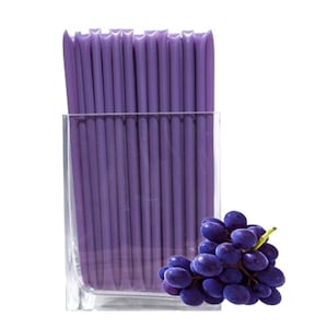 May include: A clear glass vase filled with purple wax sticks. A bunch of dark purple grapes are sitting in front of the vase.