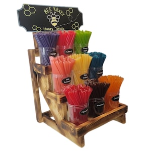 May include: A wooden display stand with multiple jars of honey sticks in various flavors, including grape, cherry, green apple, pink lemonade, cinnamon, root beer, and orange. The stand has a sign that reads "BEE KRAZY Honey Sticks".