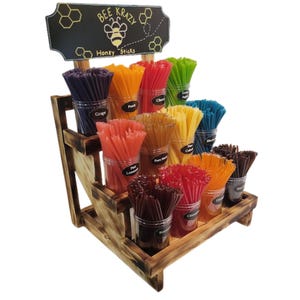 May include: A wooden tiered display stand with jars of colorful honey sticks. The jars are labeled with different flavors, including Grape, Peach, Cherry, Green Apple, Pure Honey, Pink Lemonade, Cinnamon, Raspberry, and Blueberry. The sign above the display reads "Bee Krazy Honey Sticks".