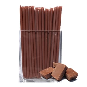 May include: A glass jar filled with brown chocolate straws.  There are several pieces of chocolate next to the jar.