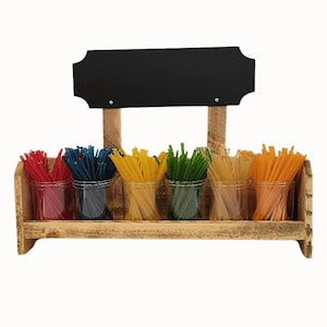 May include: A rustic wooden caddy featuring six clear plastic cups filled with thin, colorful sticks in red, blue, yellow, green, tan, and orange. Above the cups, a rectangular black chalkboard sign is mounted on two wooden support posts.