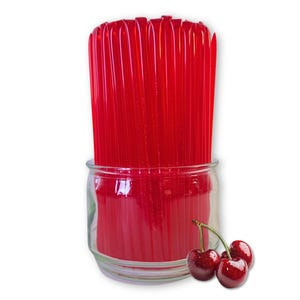 May include: A clear glass jar filled with bright red honey sticks. Three ripe cherries with green stems are in front of the jar. The honey sticks are a vibrant red color, and the cherries have a glossy finish.