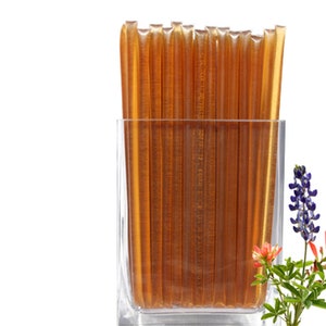 May include: A clear glass filled with honey sticks. The honey sticks are a golden brown color. The image also includes a sprig of blue and red flowers.