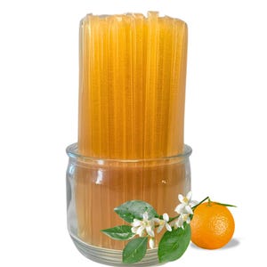 May include: A clear glass jar filled with honey sticks. The jar sits on a white surface, with an orange, white flowers, and green leaves to the side. The honey sticks are a golden yellow color.