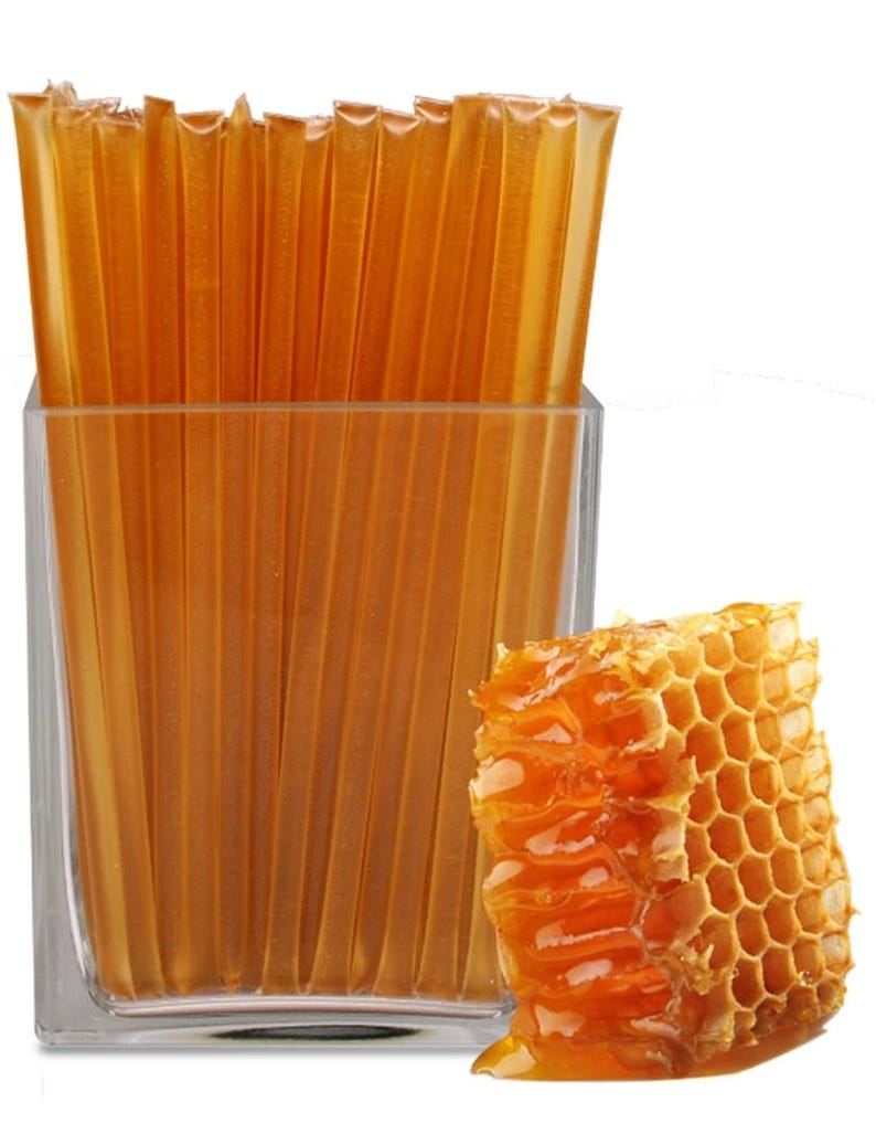 May include: A clear glass container filled with individual honey sticks, alongside a piece of honeycomb. The honey sticks are a translucent amber color, and the honeycomb is a rich, golden hue. The image is set against a white background.