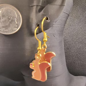 May include: Gold-colored dangle earrings featuring a cartoon squirrel design. The squirrels are brown and tan with red-brown tails, outlined in gold. The earrings are displayed on a black mannequin head.